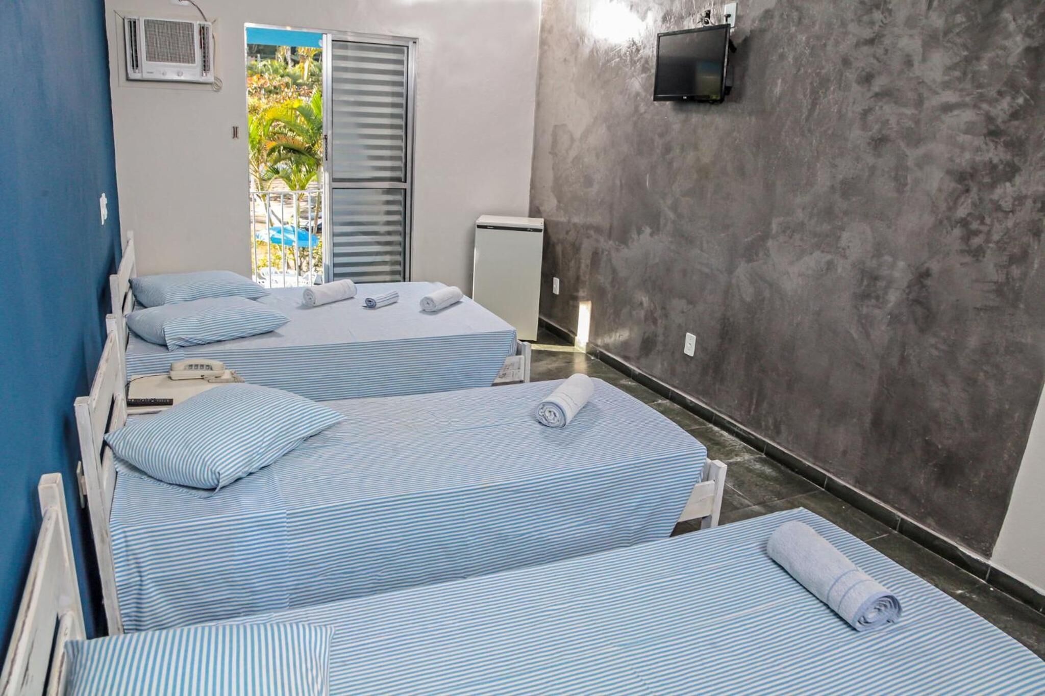 Una habitación con tres camas, cada una con ropa de cama azul a rayas y toallas enrolladas. Hay un televisor pequeño montado en la pared y una ventana con persianas.