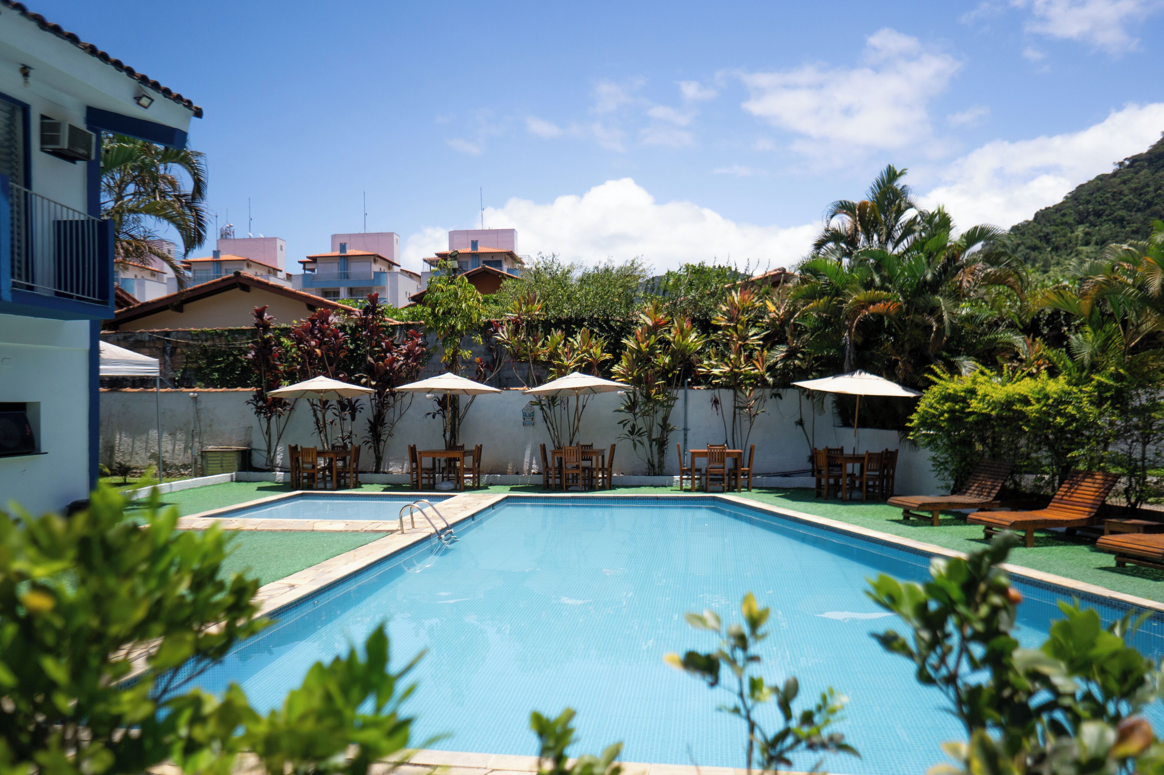 Piscina retangular com superfície de água azul cristalina, rodeada por espreguiçadeiras de madeira e guarda-sóis, com edifícios residenciais e vegetação exuberante ao fundo.