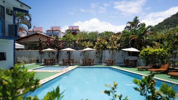 Piscina retangular com superfície de água azul cristalina, rodeada por espreguiçadeiras de madeira e guarda-sóis, com edifícios residenciais e vegetação exuberante ao fundo.