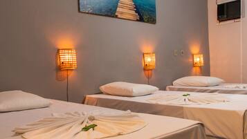 Habitación de hotel con tres camas, cada una con una almohada blanca y una toalla. Hay luces empotradas en la pared y un cuadro de un muelle.