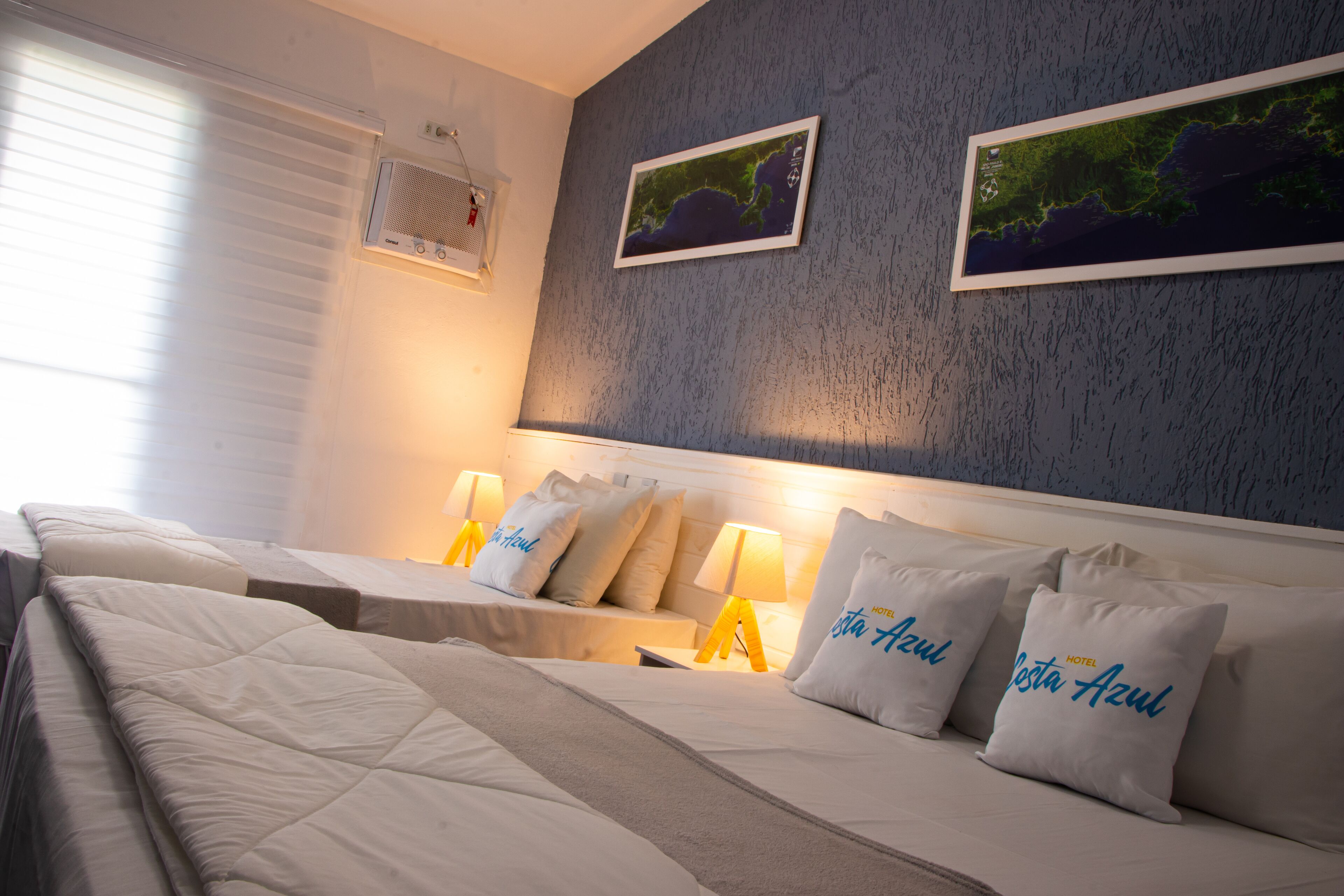 Habitación de hotel con dos camas, cada una con cabecero blanco, y dos almohadas con el nombre del hotel. Hay dos unidades de aire acondicionado montadas en la pared y dos cuadros enmarcados con paisajes.