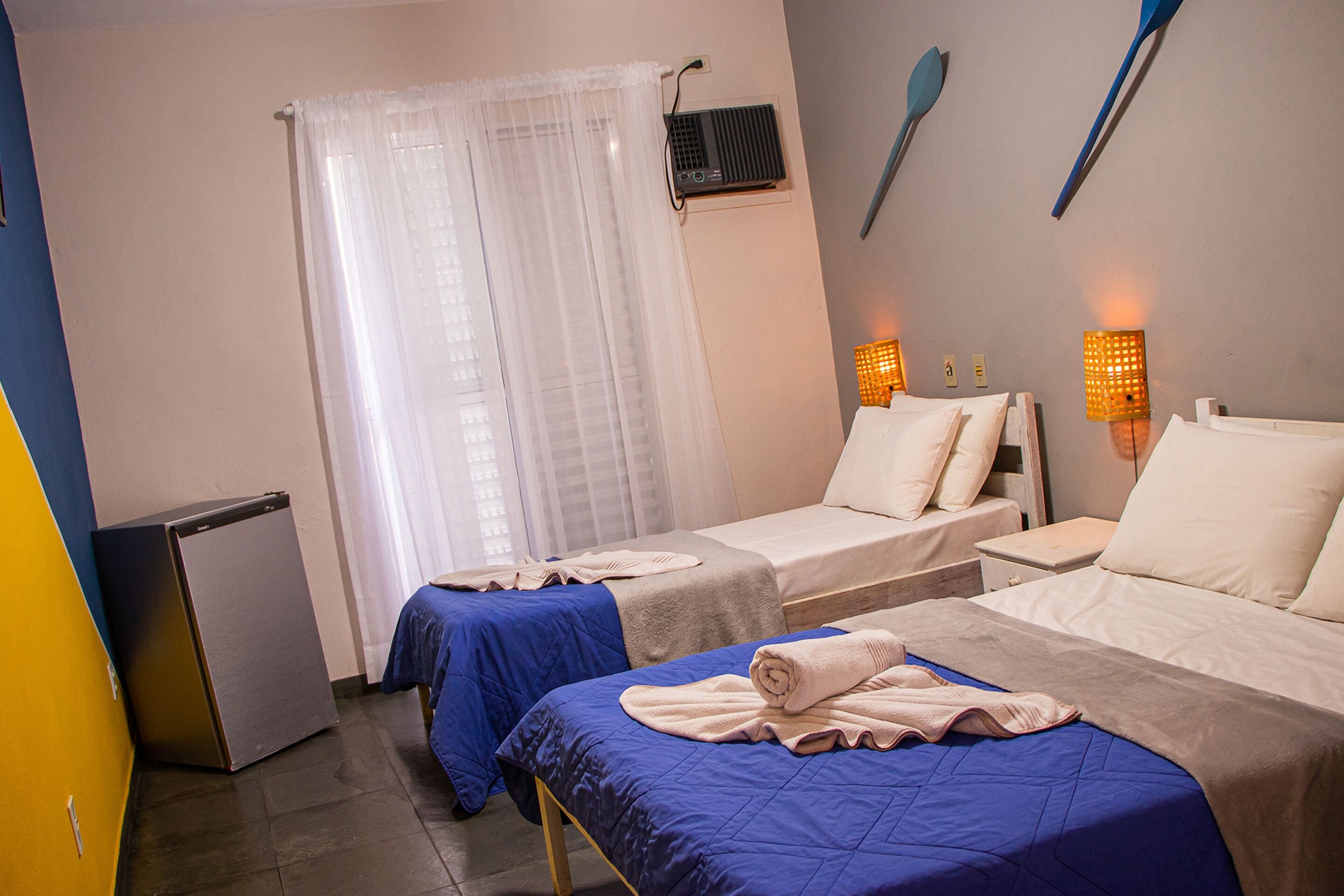 A hotel room with two beds, a small refrigerator, a window with sheer curtains, and wall-mounted lights.