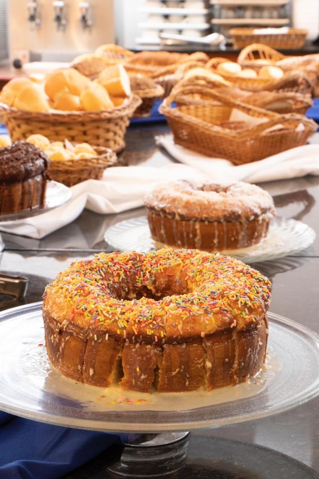 Mesa de buffet com diversos pães e doces, incluindo um bolo bundt com confeitos coloridos, muffins e cestas de pão.