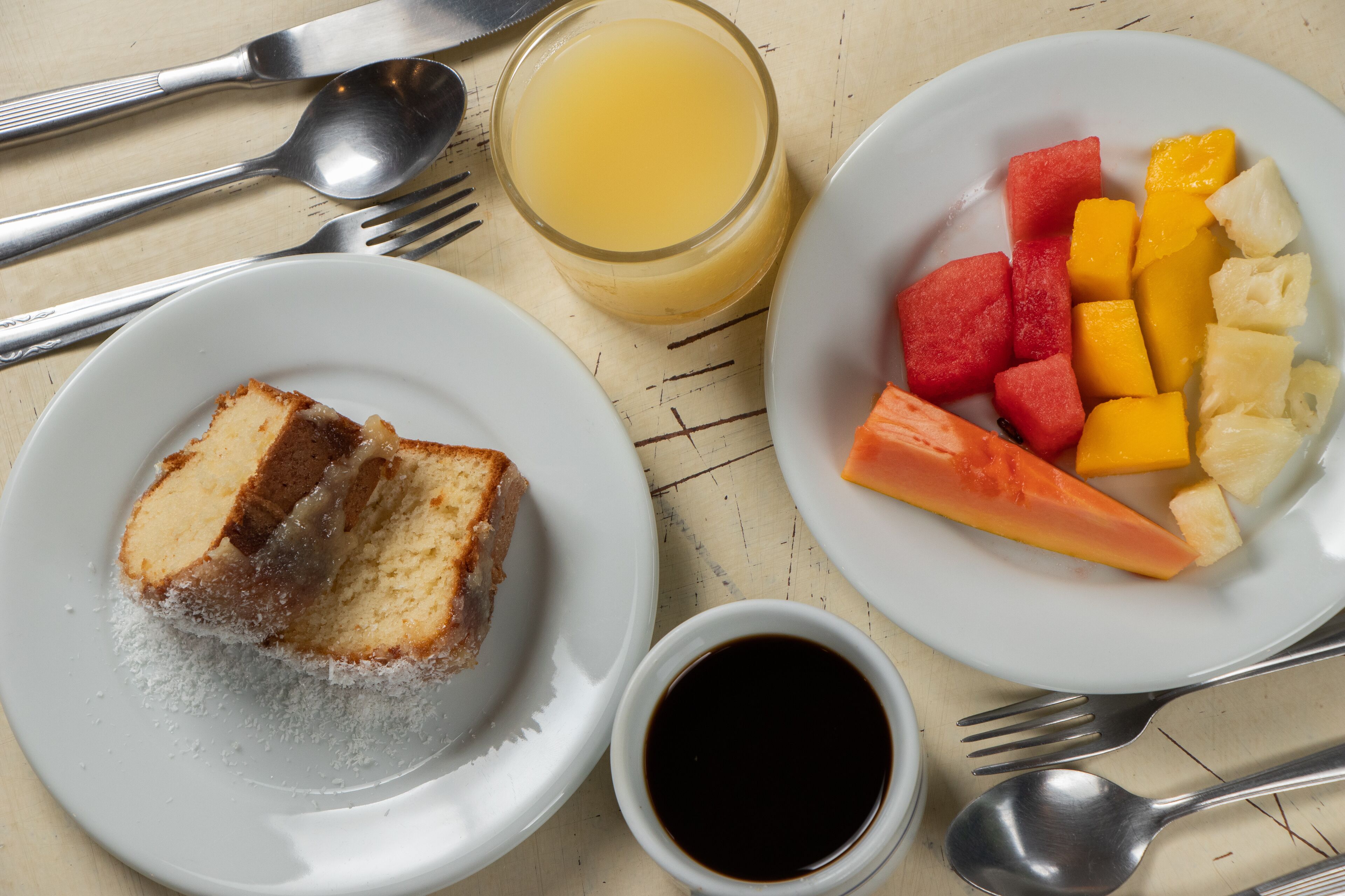 A breakfast setting with a plate of fruit, a glass of orange juice, and a slice of cake on a wooden table.