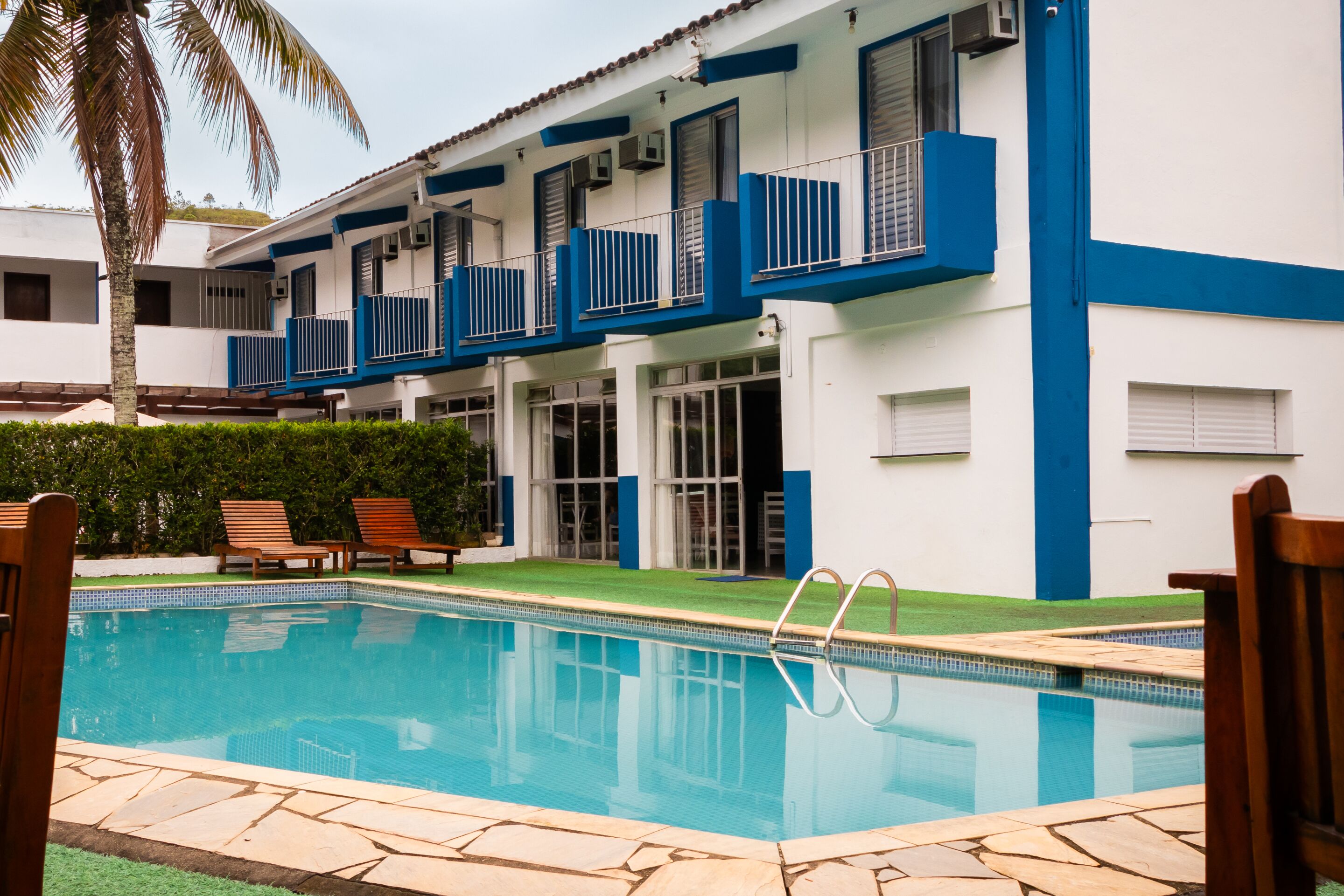 Una piscina con borde de piedra, una palmera y un edificio con paredes exteriores azules y blancas.