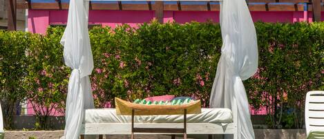 A poolside bed with a canopy, surrounded by a pool and a pink building in the background.