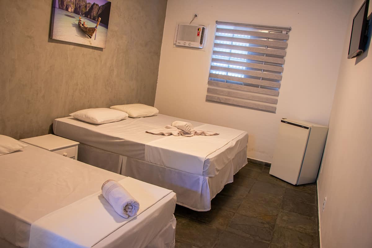 Habitación de hotel con dos camas individuales, un refrigerador pequeño, una ventana con persianas y aire acondicionado montado en la pared.
