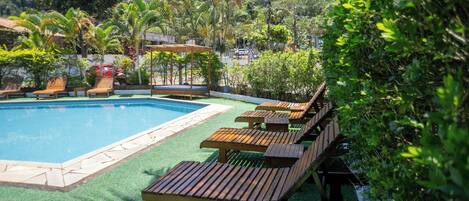 A pool area with wooden loungers and a clear blue pool surrounded by greenery and a hilly backdrop.