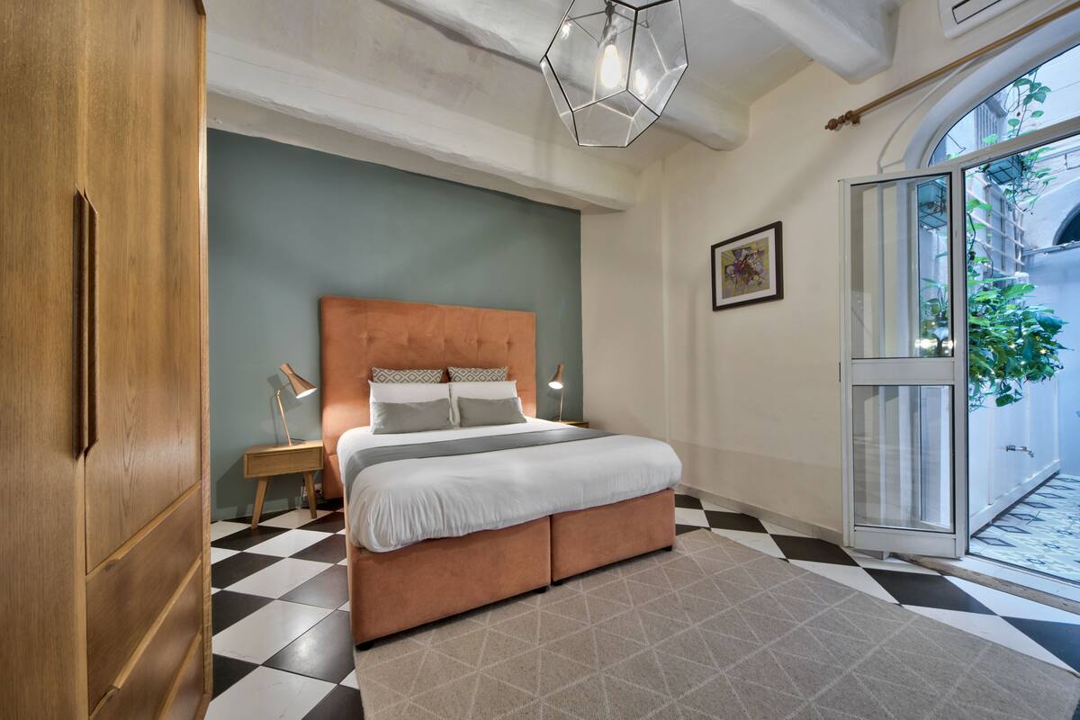 A bedroom with a large bed, a wooden wardrobe, a desk with a lamp, and a view of the outside through a glass door.