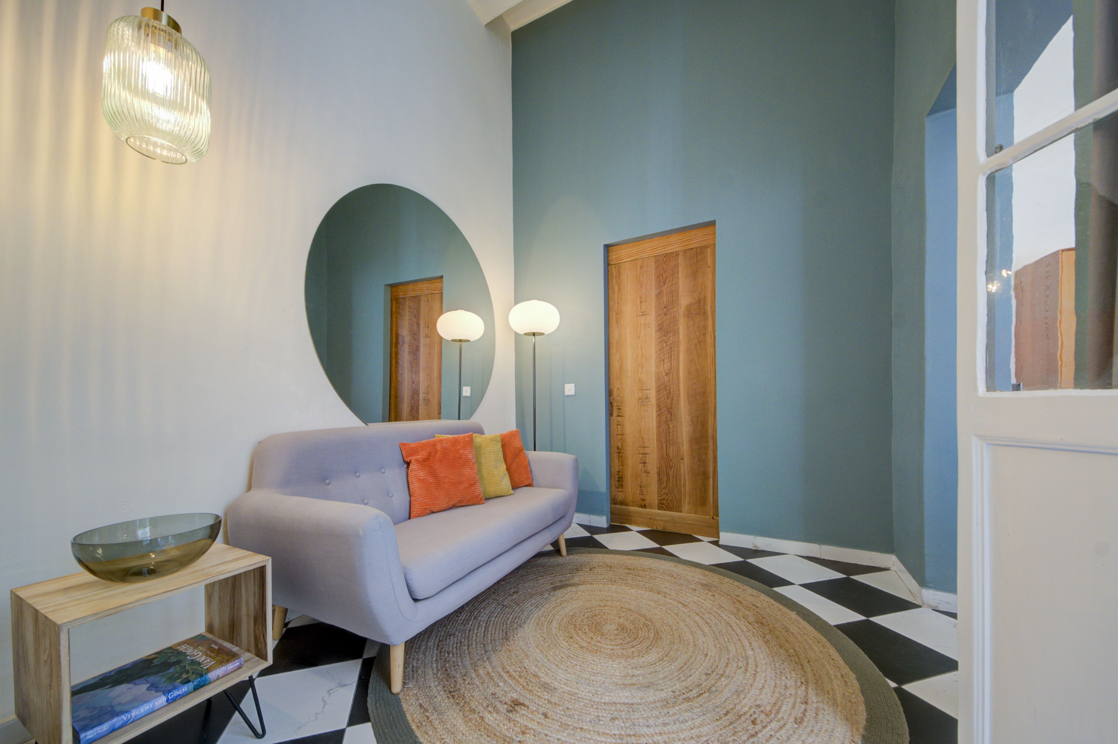 Ein Zimmer mit einem hellvioletten Sofa, einem Holznachttisch mit einer Glasbowl, einem gemusterten Teppich und einem karierten Boden.
