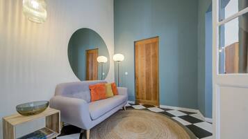 Ein Zimmer mit einem hellvioletten Sofa, einem Holznachttisch mit einer Glasbowl, einem gemusterten Teppich und einem karierten Boden.