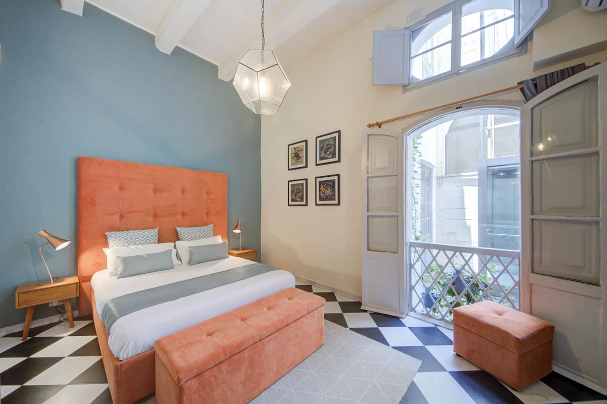 A bedroom with a checkered floor, a large bed with an orange headboard, a bench, and a window with a view.