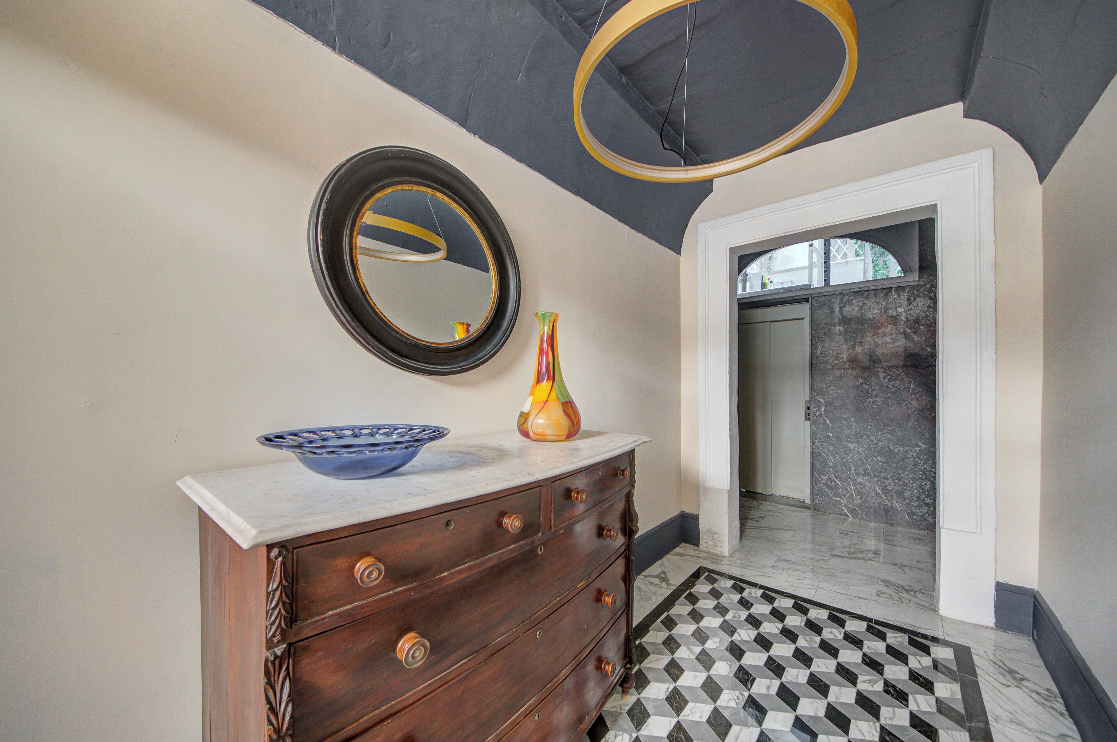 Une salle de bain avec un sol en marbre, une commode en bois, un bol bleu, un vase et un miroir.