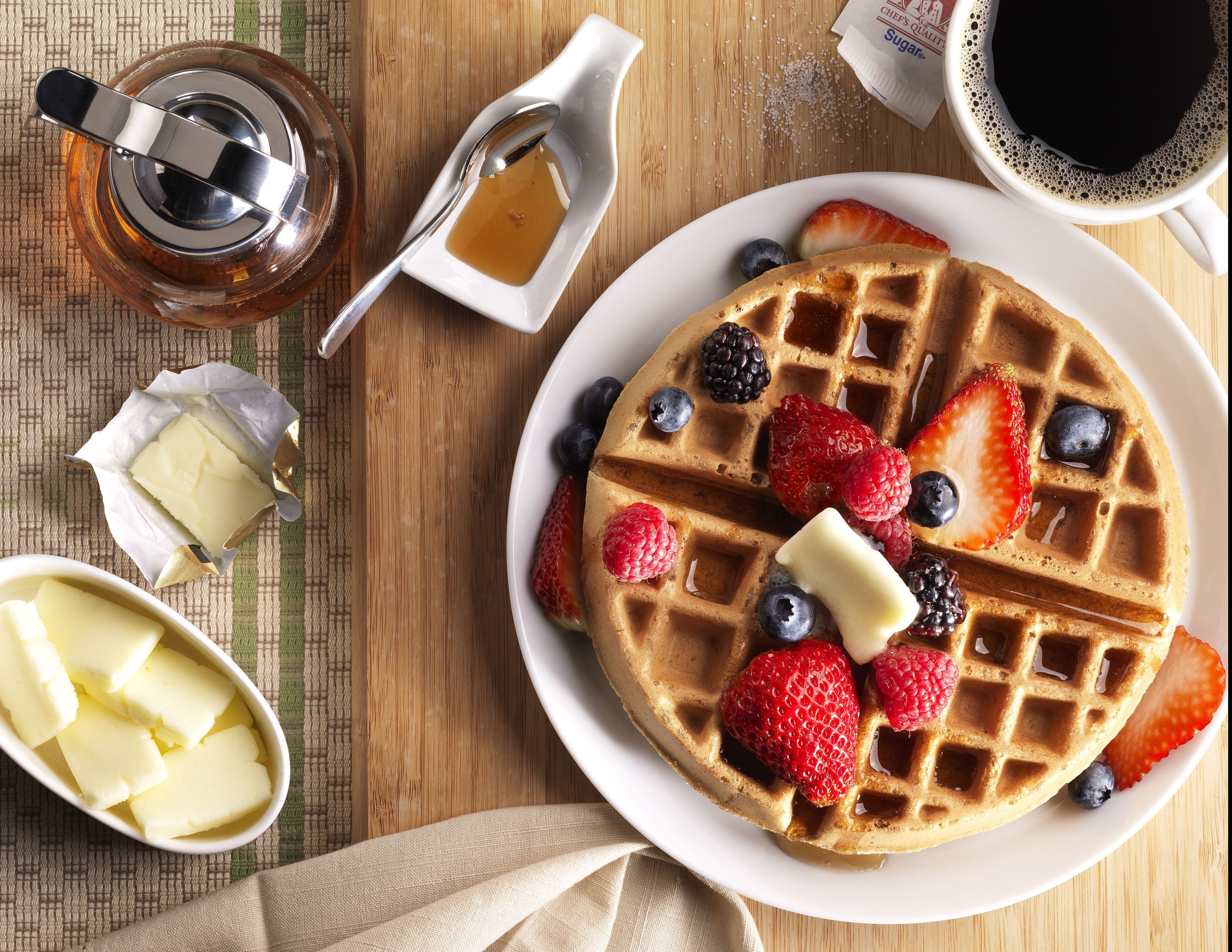 Ein Teller mit Waffeln, Beeren und Butter, ein Glas Sirup und eine Tasse Kaffee auf einem Holztisch.
