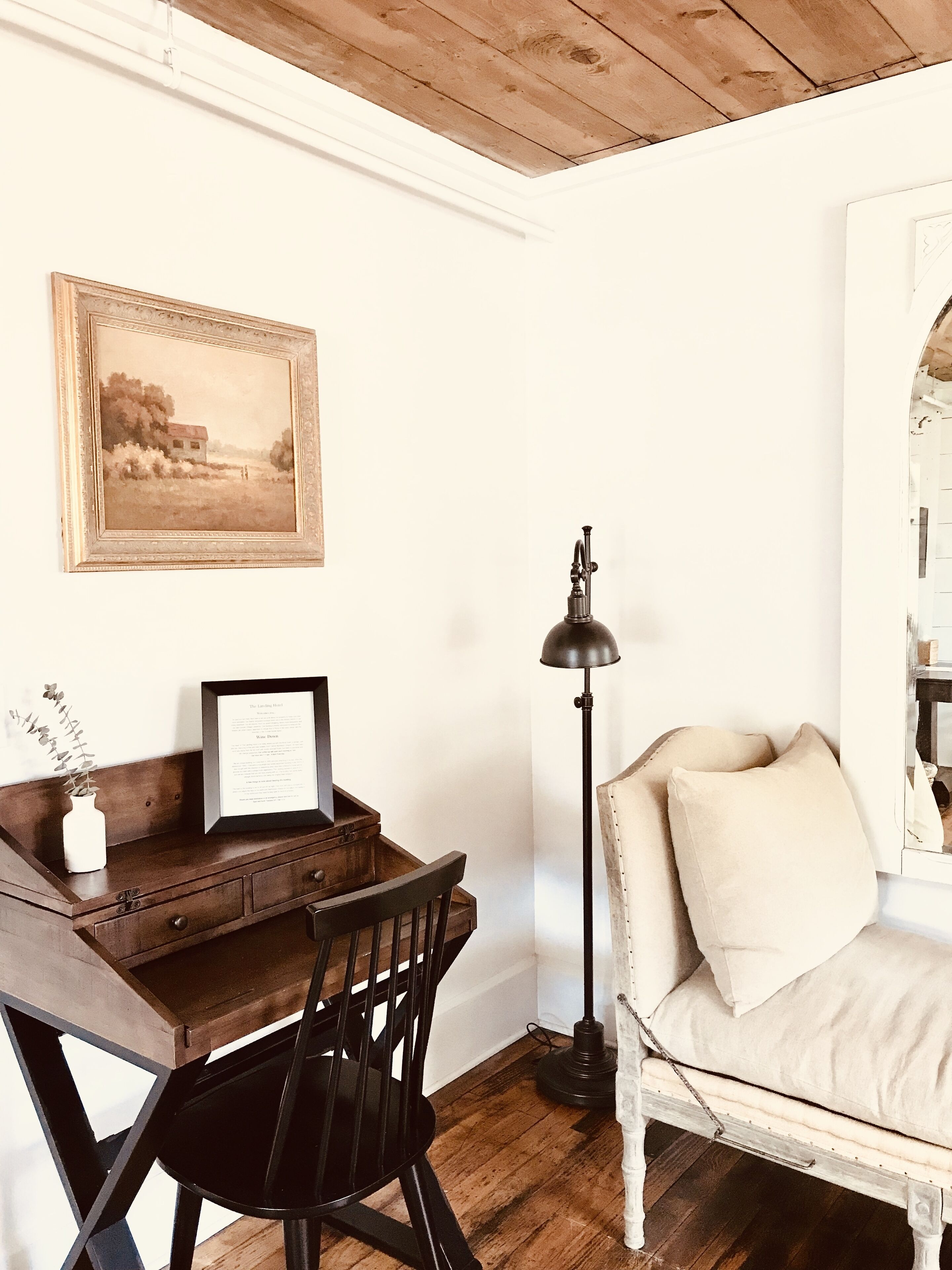 A cozy room with a wooden desk, a framed picture, a black chair, a floor lamp, and a white sofa with cushions.