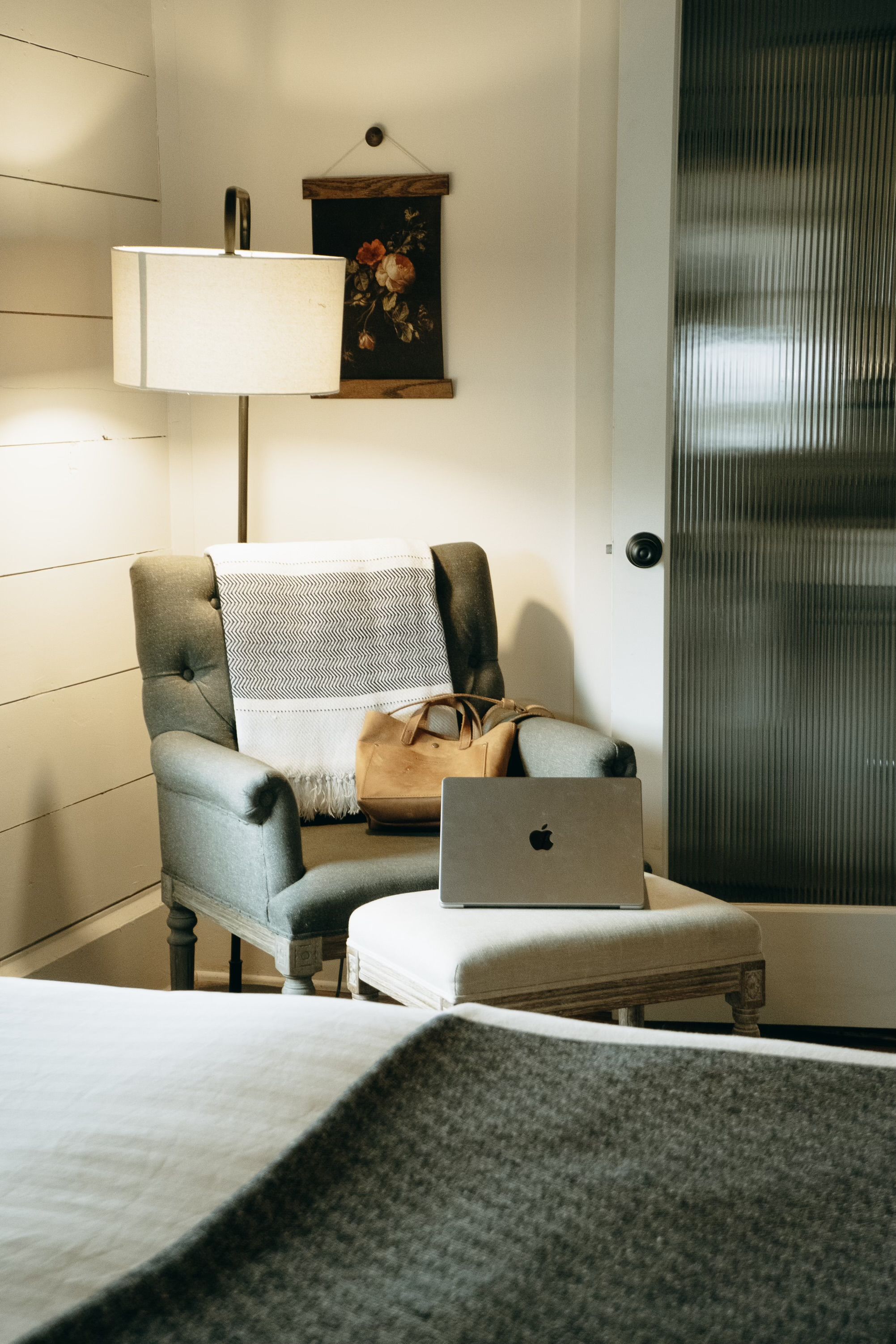 Una habitación de hotel con una cama, una silla con una computadora portátil, una lámpara y un cuadro en la pared.