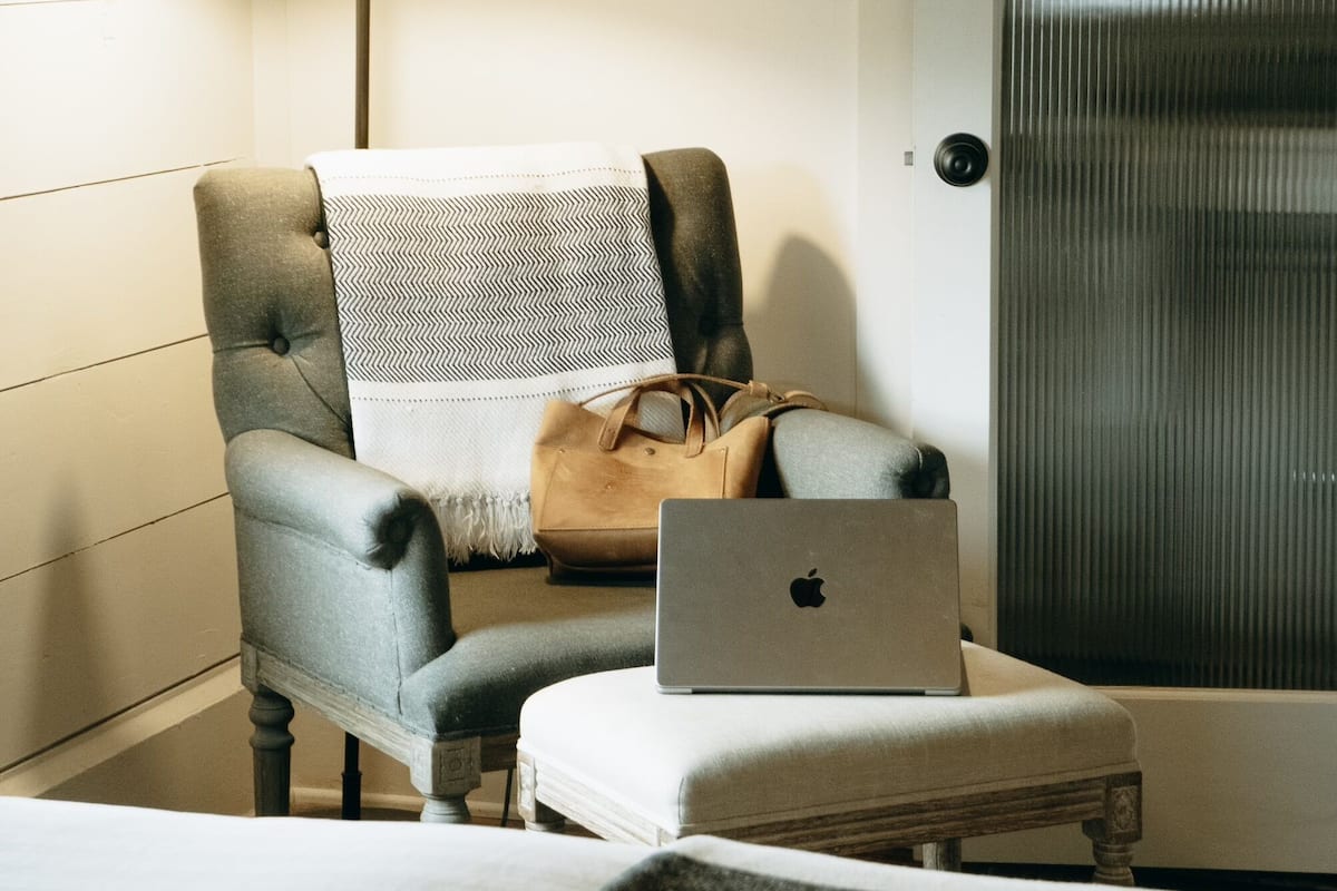 A hotel room with a grey upholstered armchair, a small white table, a laptop, and a handbag on the chair.