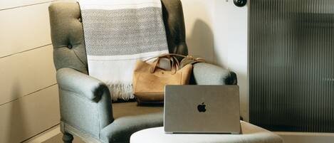 Habitación de hotel con un sillón tapizado en gris, una mesita pequeña, una computadora portátil y un bolso sobre el sillón.