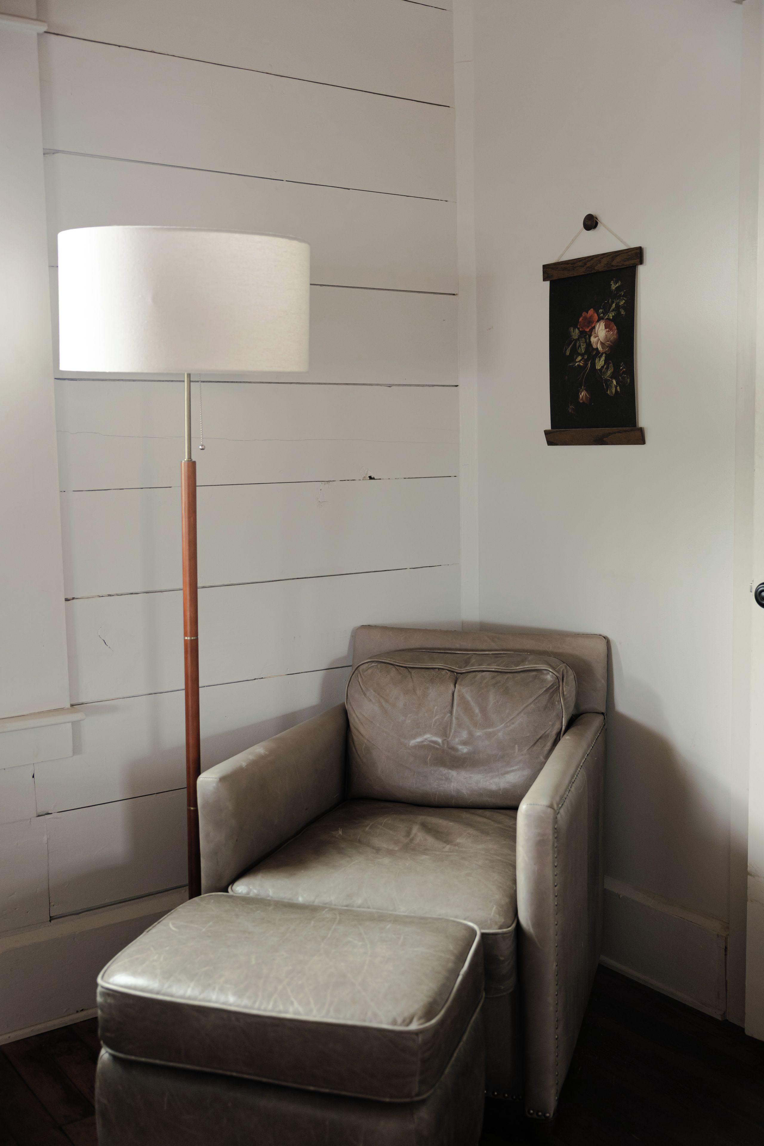 A corner room with a chair and ottoman, a floor lamp, and a framed picture on the wall.