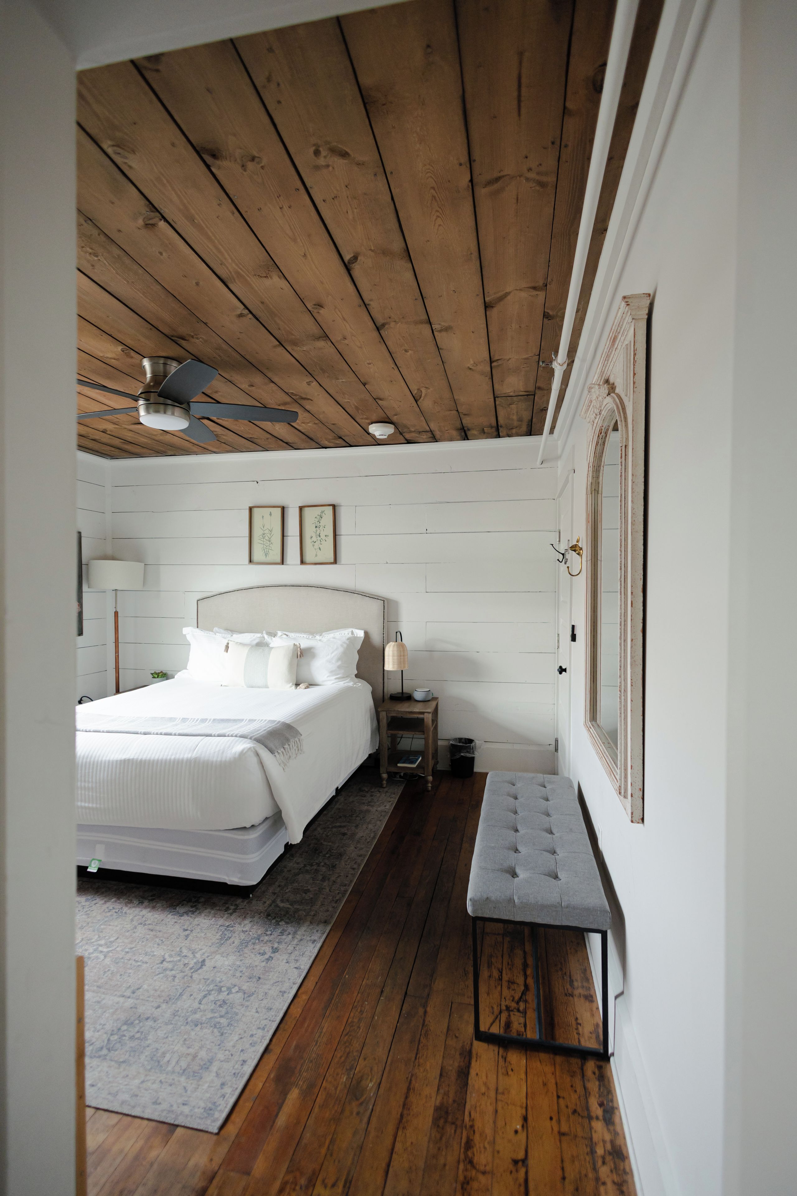 Ein Schlafzimmer mit Holzboden, einem Bett mit weißer Bettwäsche, einem Nachttisch, einer Bank und einem gerahmten Bild an der Wand.