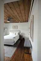 Un dormitorio con techo de madera, cama con ropa blanca, mesita de noche, un banco y un cuadro en la pared.