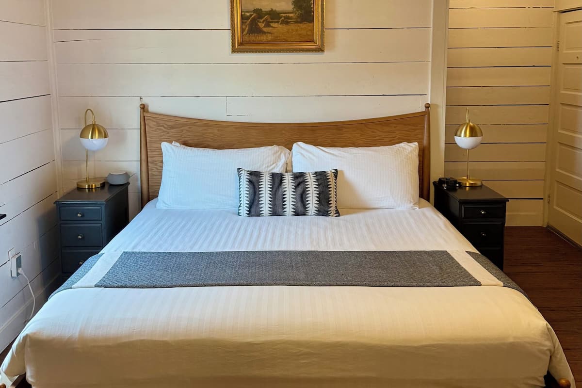 A wooden-paneled bedroom with a large bed, two bedside tables with lamps, a hanging light fixture, and a framed picture on the wall.
