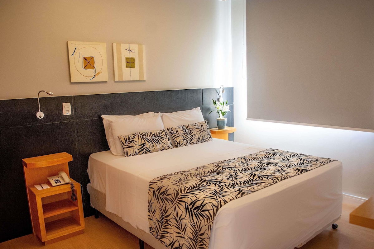 Una habitación de hotel con una cama, una mesita de noche con un teléfono, una lámpara de pared y dos obras de arte enmarcadas.