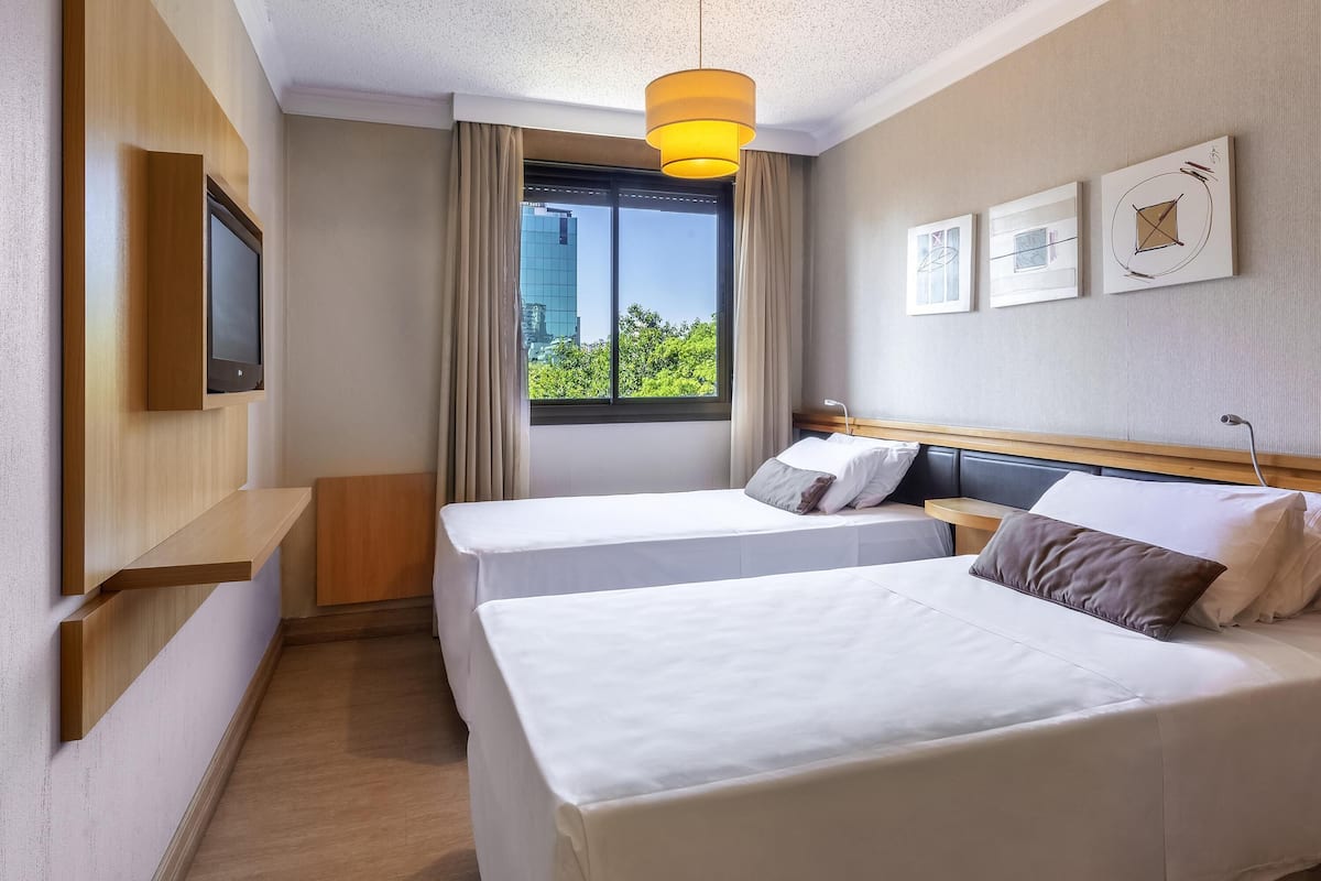 Habitación de hotel con dos camas, un televisor fijado a la pared, una ventana con cortinas y obras de arte en la pared.