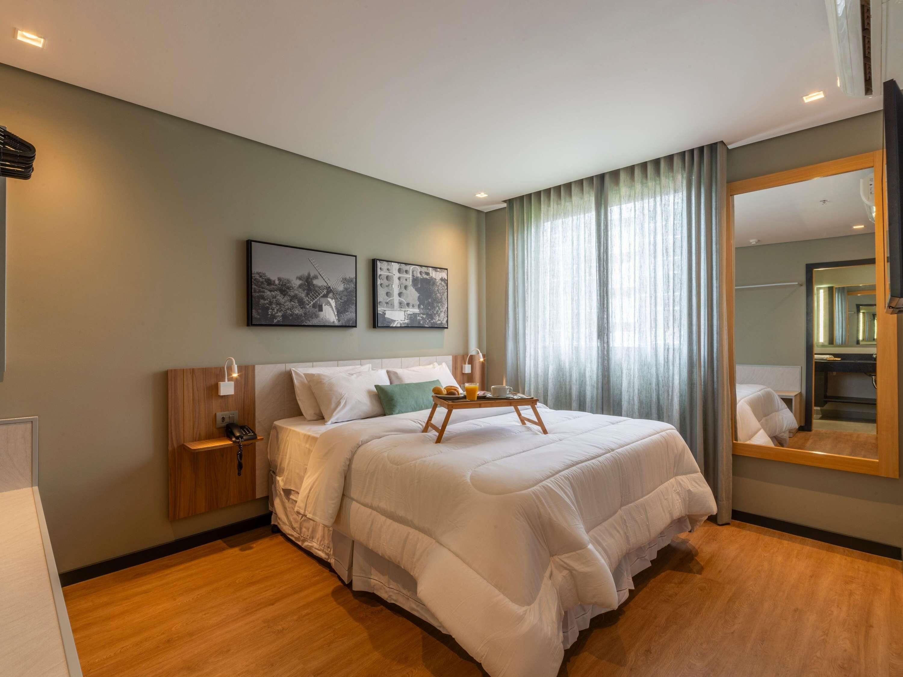 Habitación de hotel con una cama grande, mesitas de noche, una lámpara de noche, una bandeja con alimentos para el desayuno y un espejo amplio.
