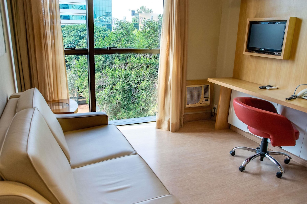 Una habitación de hotel moderna con sofá, una silla de escritorio roja, un escritorio con televisor de pantalla plana y un ventanal con vista a la ciudad.