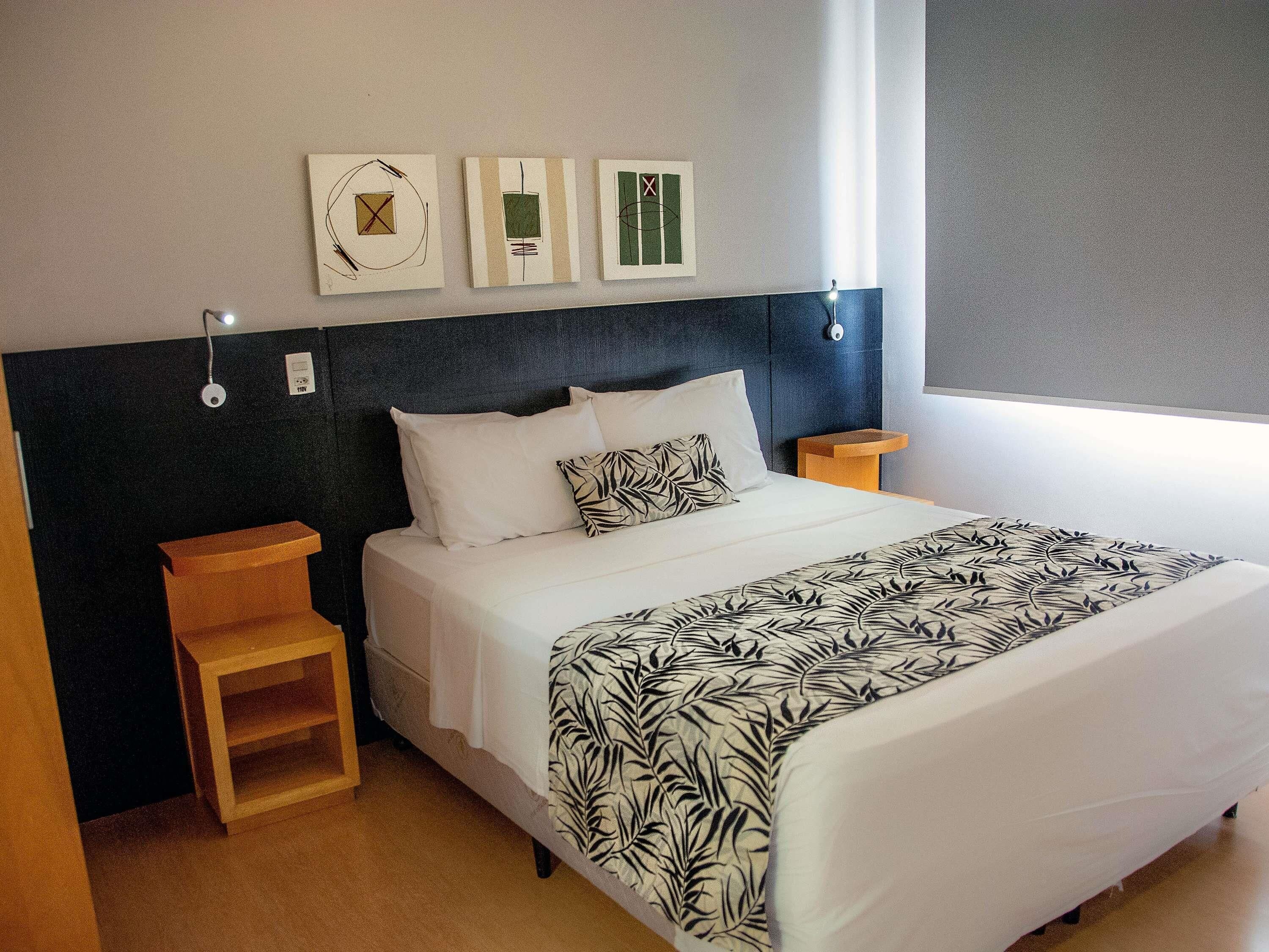 Uma cama bem arrumada com colcha estampada, cabeceira escura e três quadros na parede.