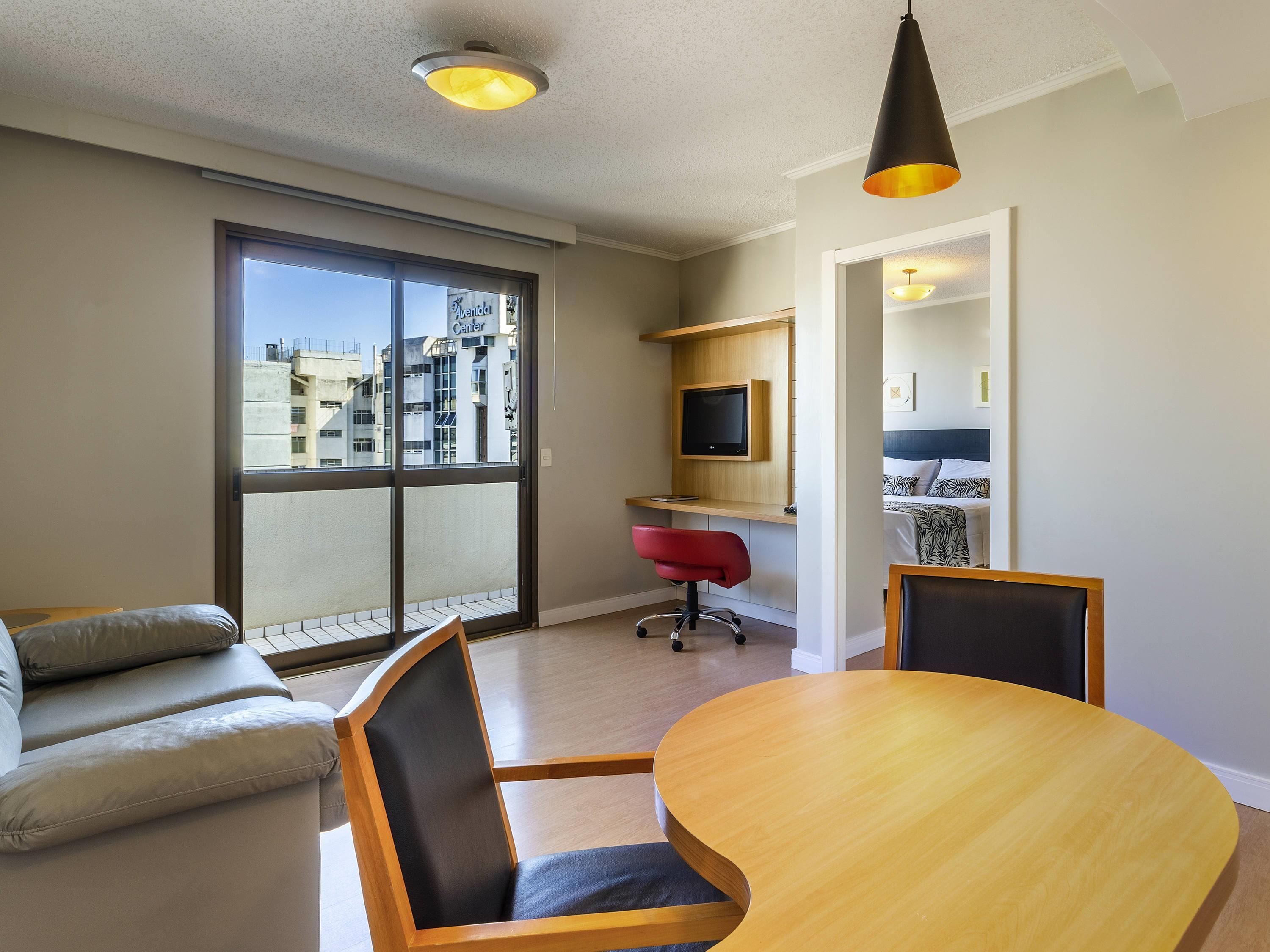 A modern living room with a large window, a sofa, a dining table, a TV, and a red office chair.