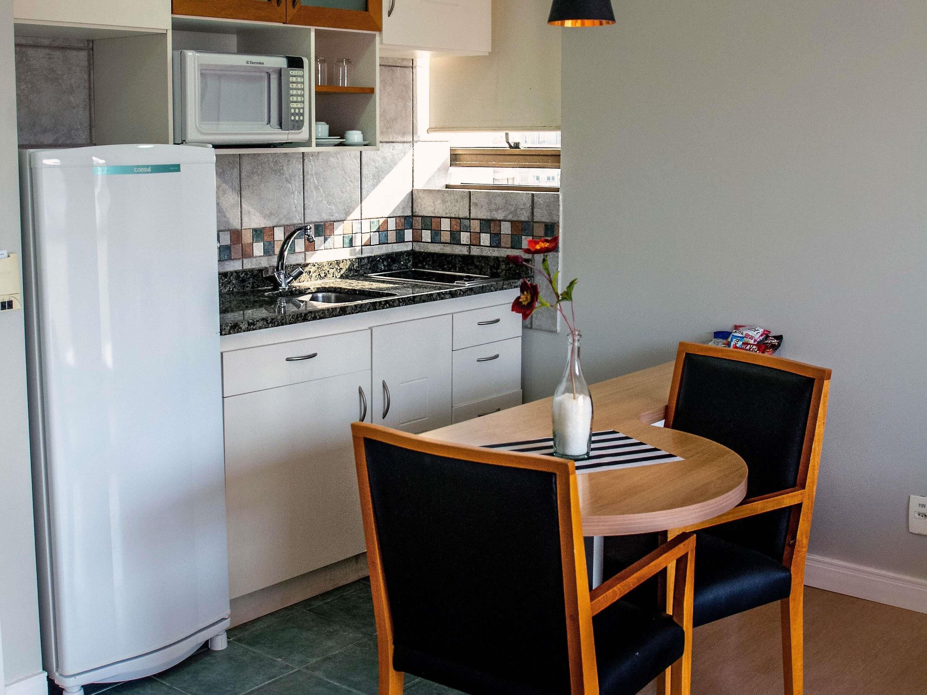 Cozinha compacta com geladeira, micro-ondas e pia. Há uma mesa de jantar com duas cadeiras e um vaso com flores.
