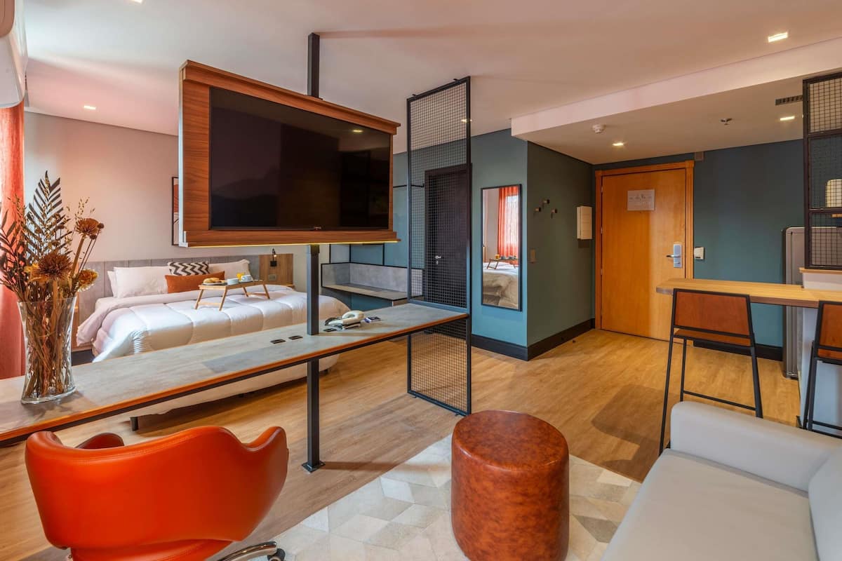 Una habitación de hotel moderna con cama, mesita de noche, televisor y un área de descanso con sillón y reposapiés.