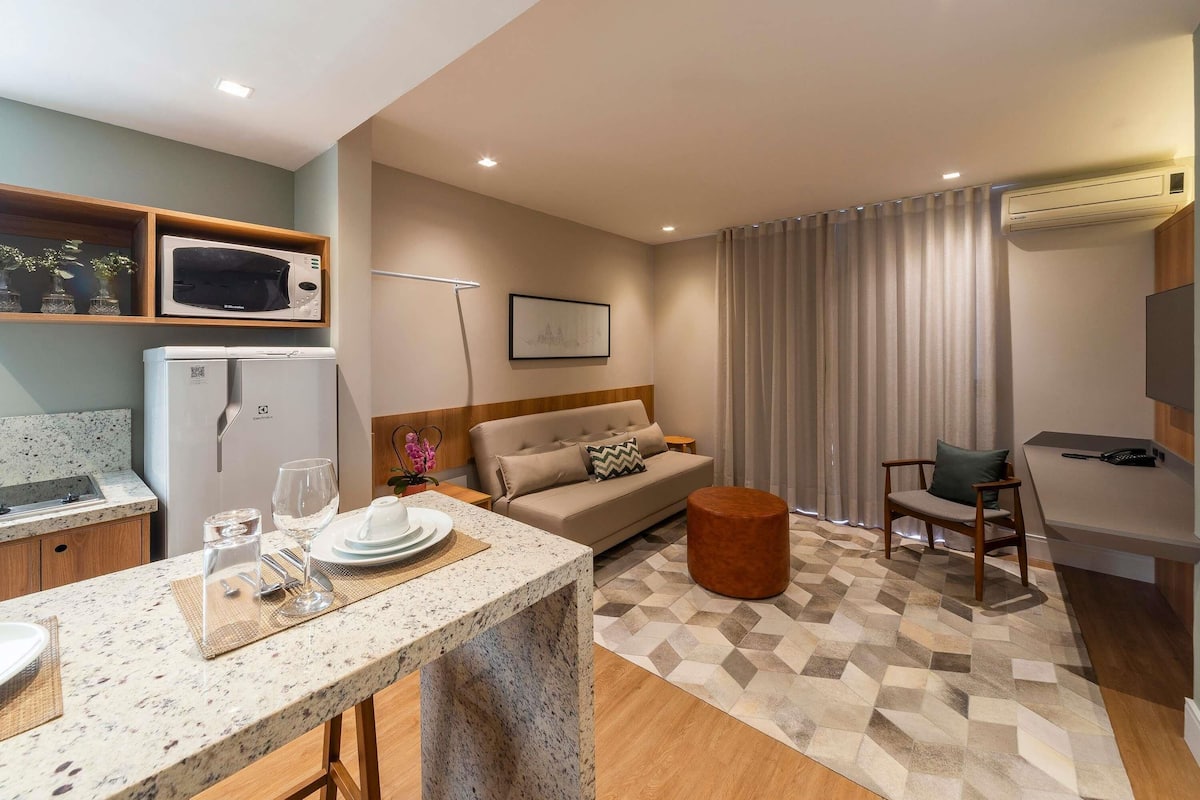 Una habitación de hotel moderna con kitchenette, zona de comedor, sofá, otomán pequeño y redondo, y escritorio con silla.