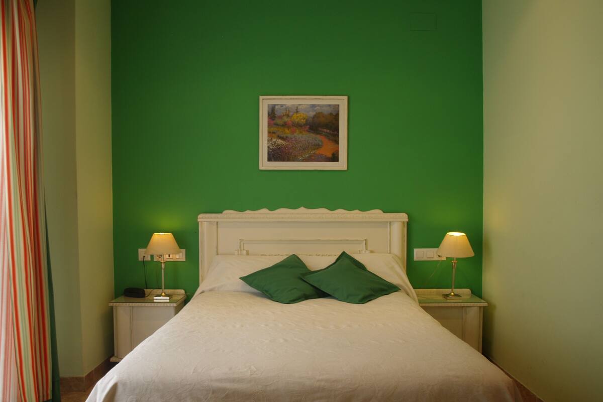 Un llit ben arrenjat amb roba de llit blanca i dos coixins verds, flanquejat per dues taules de nit amb làmpades, en una habitació amb parets verdes i un quadre encimbellat sobre el llit.