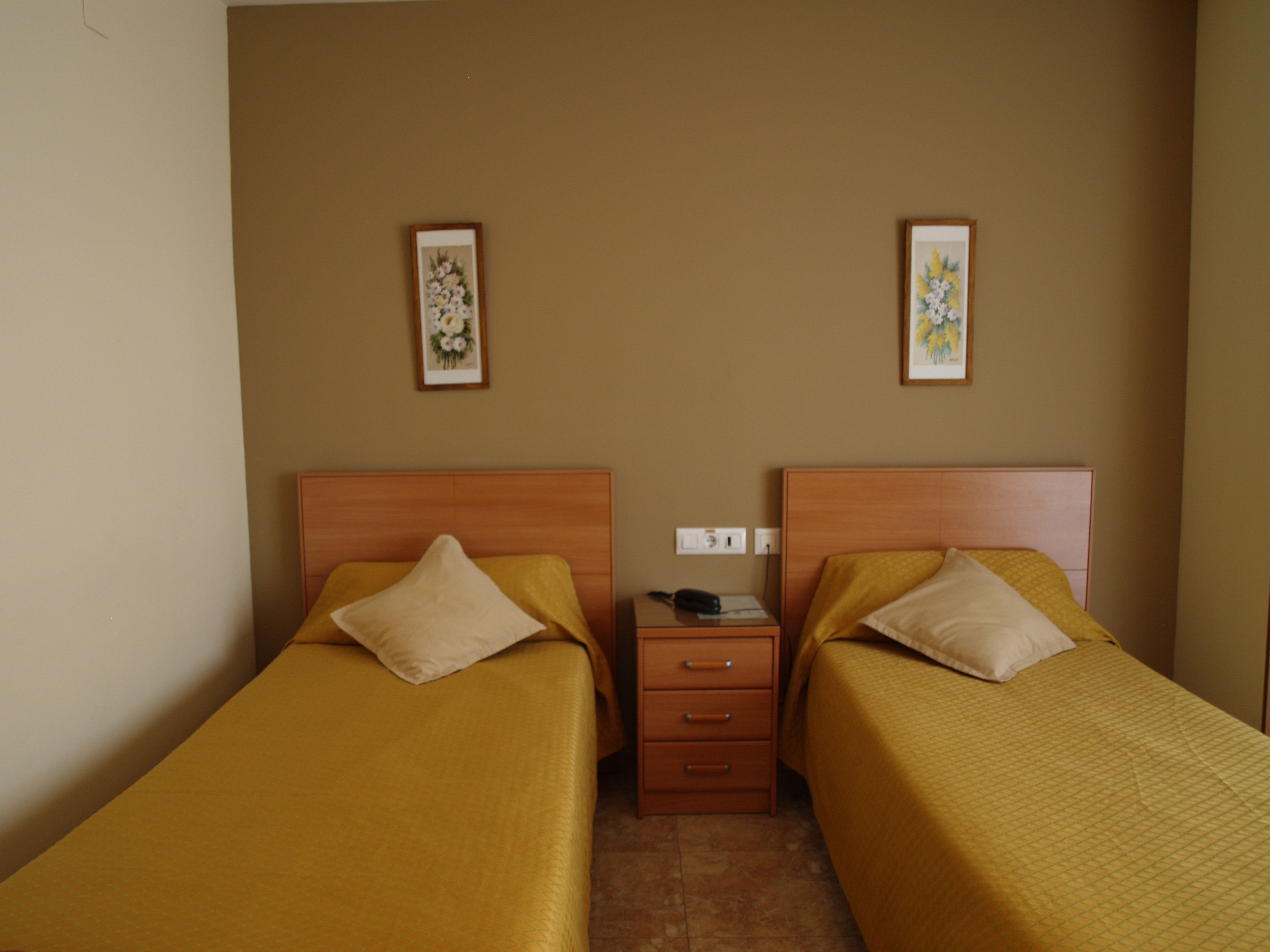 A hotel room with two single beds, each with a wooden headboard and a beige pillow. There is a wooden nightstand between the beds with a lamp and a phone. The walls are beige, and there are two framed floral pictures on the wall.