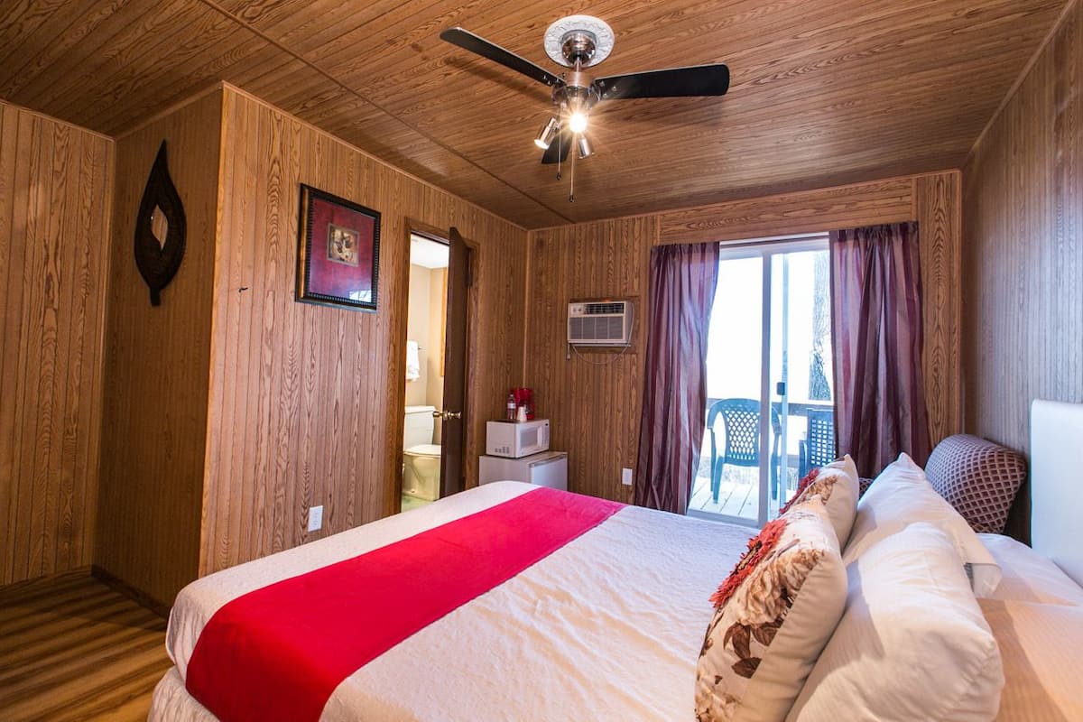 A wooden-paneled room with a bed, a ceiling fan, a window with curtains, and a small table with a microwave.