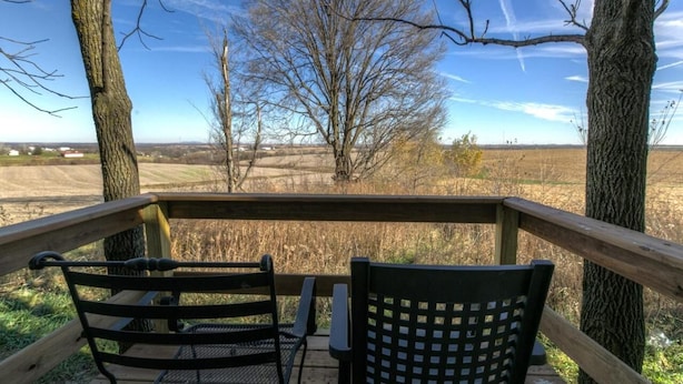 Un terrazza in legno con due sedie in metallo e una ringhiera in legno che si affaccia su un paesaggio rurale con alberi e campi aperti.