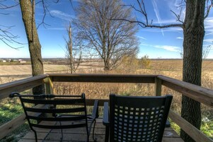 Una terraza de madera con dos sillas de metal y baranda de madera que da a un paisaje rural con árboles y campos abiertos.