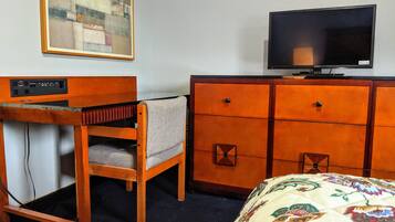 Una camera d'hotel con una scrivania in legno, un televisore su un comò, un letto con copripiumino floreale e un quadro appeso al muro.