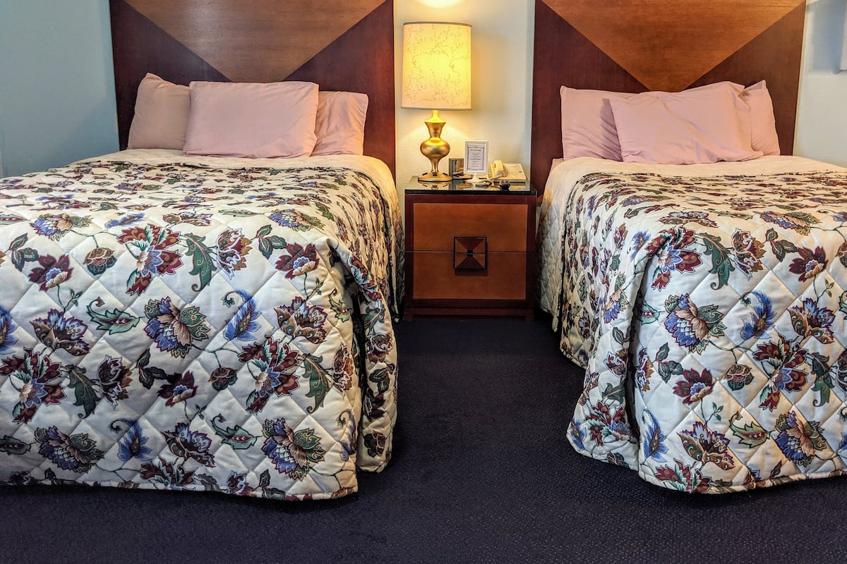 A hotel room with two beds, each with floral-patterned quilts, a nightstand with a lamp, and a wooden headboard.