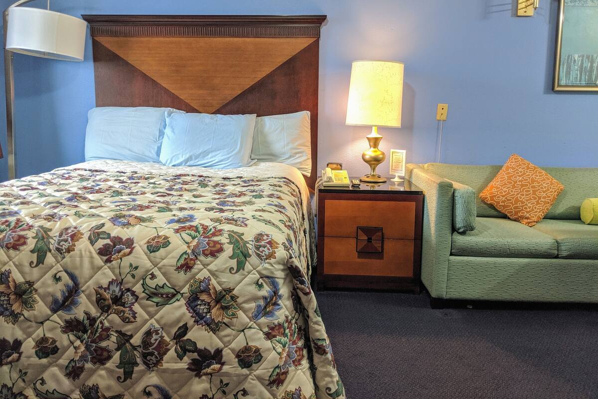 A hotel room with a bed, a nightstand, a lamp, a green sofa, and a wall clock.