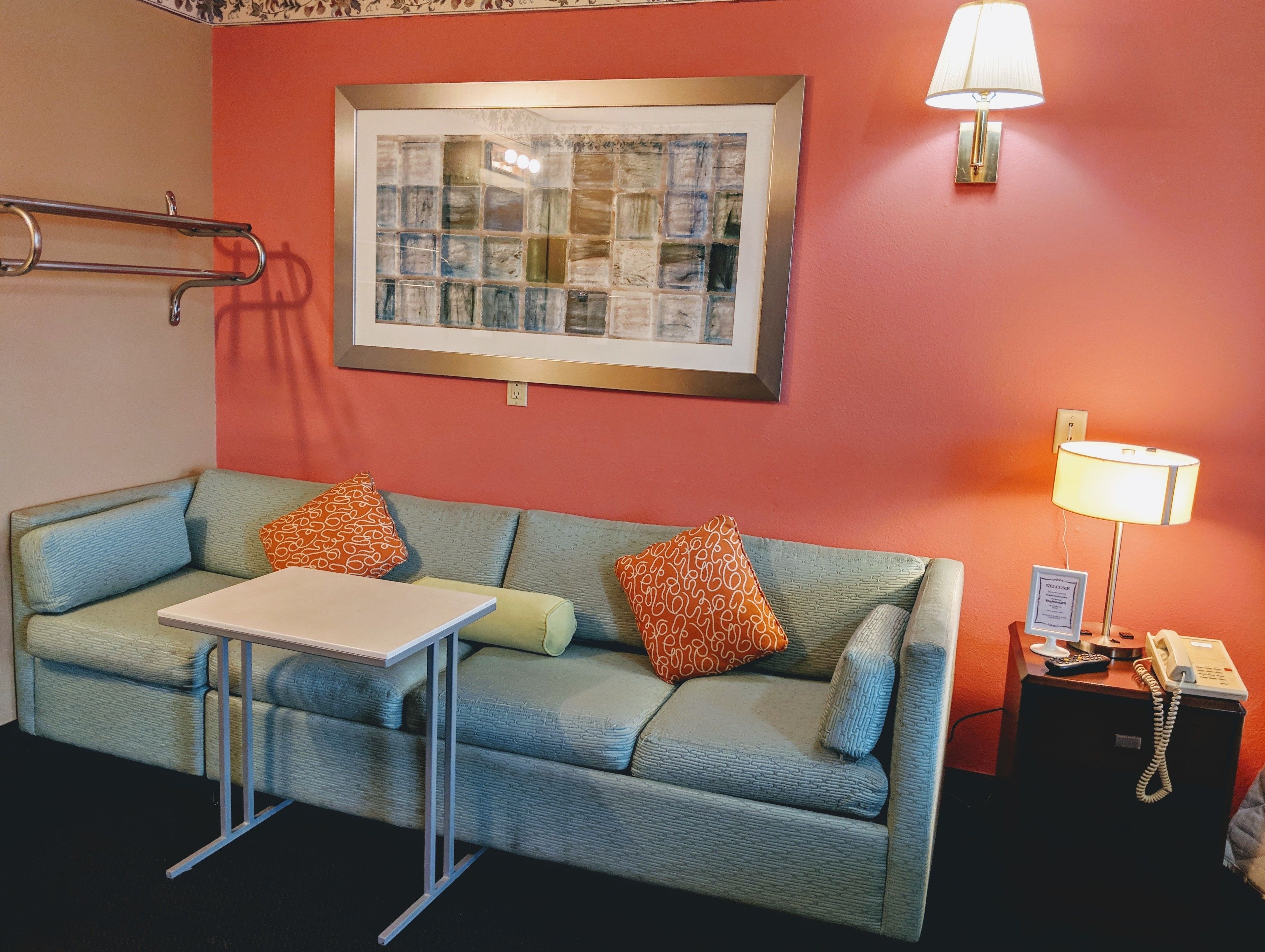 A room with a blue sofa, two pillows, a small table, a framed picture on the wall, a lamp, and a phone on a side table.