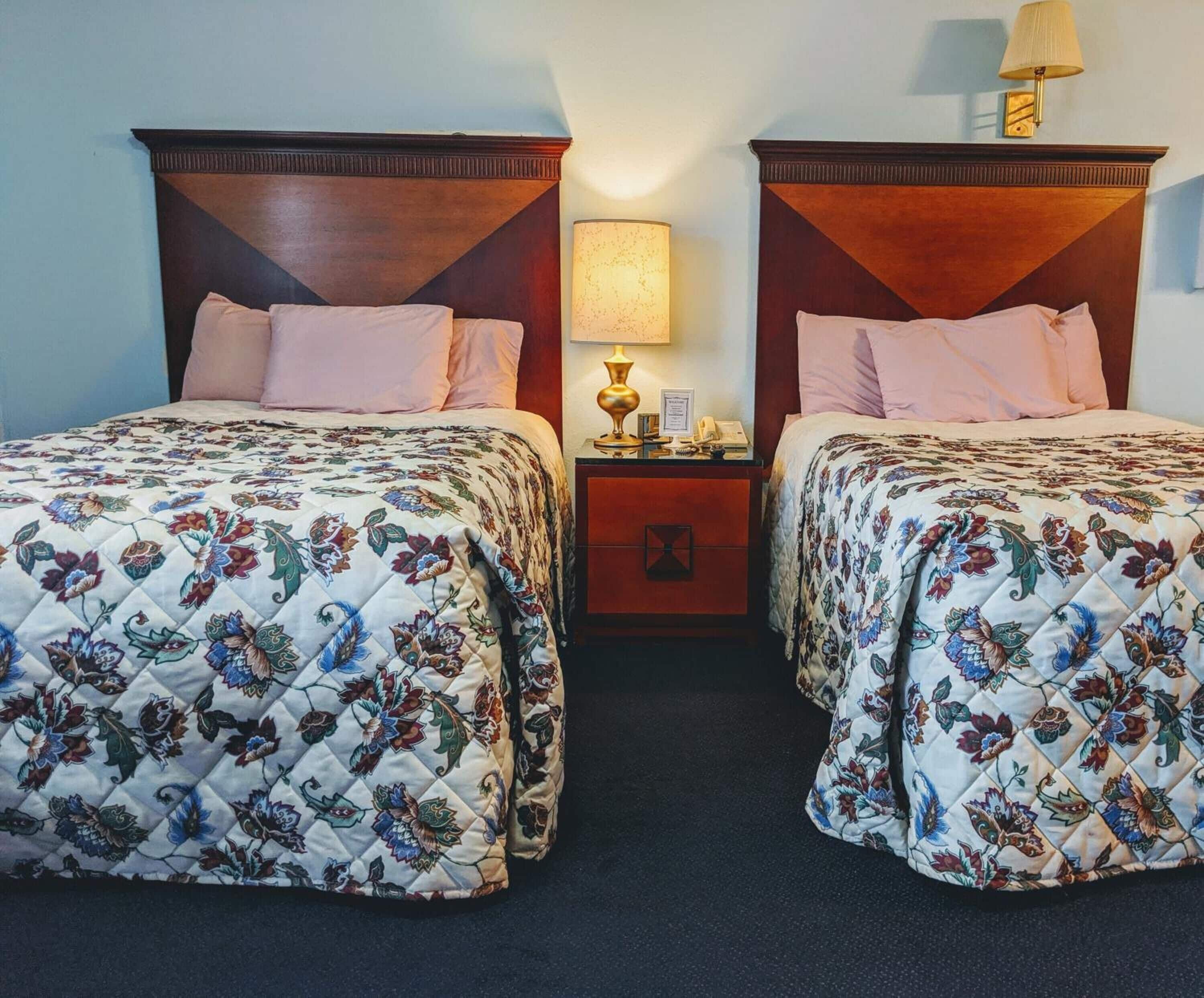Ein Hotelzimmer mit zwei Betten, jeweils mit einem floralen gesteppten Bettüberwurf und passenden Kopfteilen. Zwischen den Betten befindet sich ein Nachttisch mit einer Lampe und einem Telefon.