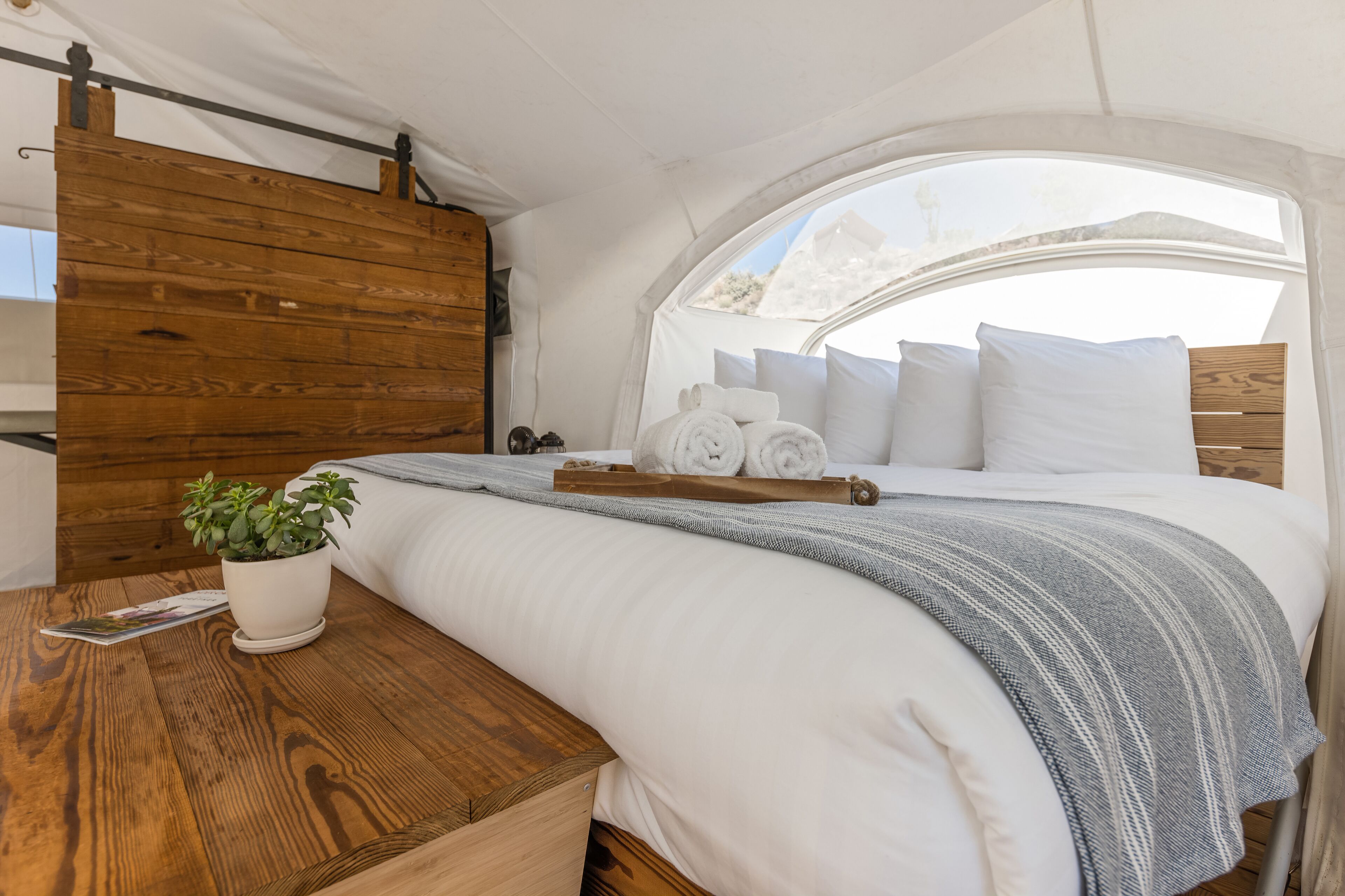 A cozy bedroom with a wooden headboard, a neatly made bed with white linens, a grey striped throw, and a wooden nightstand with a potted plant and a magazine.