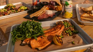 A meal setup with various dishes on trays, including salmon, greens, and bread, with a can of San Pellegrino and a bottle of lemon juice.
