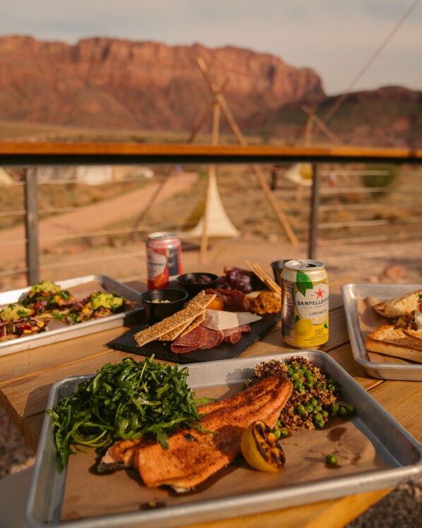 A meal setup with various dishes on trays, including salmon, greens, and bread, with a can of San Pellegrino and a bottle of lemon juice.