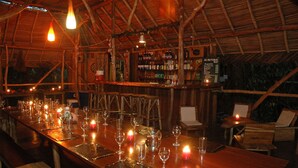 Un bar rustique en bois, doté d’un toit de chaume, où l’on trouve une variété de bouteilles et de verres, ainsi qu’un tableau affichant le menu.
