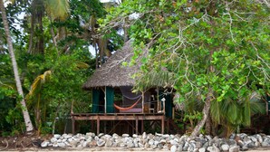 Un bungalow au toit de chaume, doté d’un hamac, entouré d’une côte rocheuse et d’une végétation tropicale luxuriante.